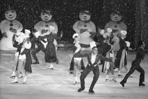 Eine Gruppe von Menschen in Kostümen fährt auf einer Eisbahn im Schnee, mit Schneemännern im Hintergrund, in Schwarzweiß abgebildet.