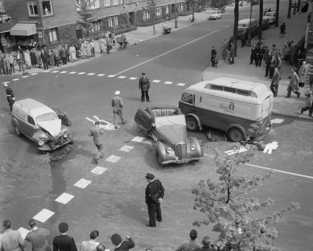 Schwarzes und weißes Bild eines Autounfalls an einer Straßenkreuzung mit mehreren Fahrzeugen, darunter ein Lieferwagen, und einer Gruppe von Menschen, die daneben stehen, mit Gebäuden, Bäumen, Pfählen und Texttafeln im Hintergrund.