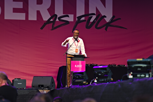 Ein Mann in einem weißen Hemd und braunen Hosen steht auf einer Bühne und hält ein Mikrofon in der Hand, mit einem pinken Banner im Hintergrund, auf dem 'Berlin as Fuck' steht, und Lautsprechern auf der Bühne.