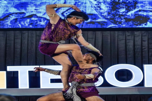 Zwei Performer in violetten Kleidern führen einen akrobatischen Hebemanöver auf der Bühne vor einem Publikum aus, mit schwarzem Vorhang und Bildschirmhintergrund.