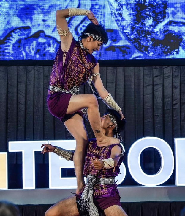 Zwei Performer in violetten Kleidern führen einen akrobatischen Hebemanöver auf der Bühne vor einem Publikum aus, mit schwarzem Vorhang und Bildschirmhintergrund.