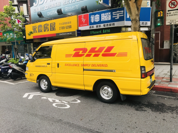 Gelbes DHL-Lieferfahrzeug auf Straße neben Gebäuden, Bäumen, Schildern, Mästen, Ampeln und einer Person auf dem Gehweg.