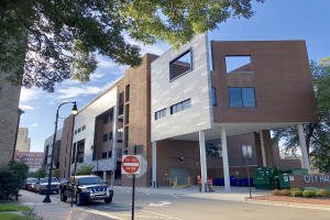Großes Rathausgebäude mit zahlreichen Fenstern, umgeben von Straßeninfrastruktur einschließlich Masten, Laternen, Fahrzeugen, Schildern und Verkehrselementen, mit Bäumen und einem bewölkten Himmel im Hintergrund.