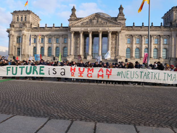 Eine Gruppe von Menschen hält ein Transparent mit der Aufschrift "Zukunft ist ein Mensch" vor dem Reichstaggebäude in Berlin, Deutschland, mit sichtbaren architektonischen Details und Flaggen im Hintergrund bei einem bewölkten Himmel.