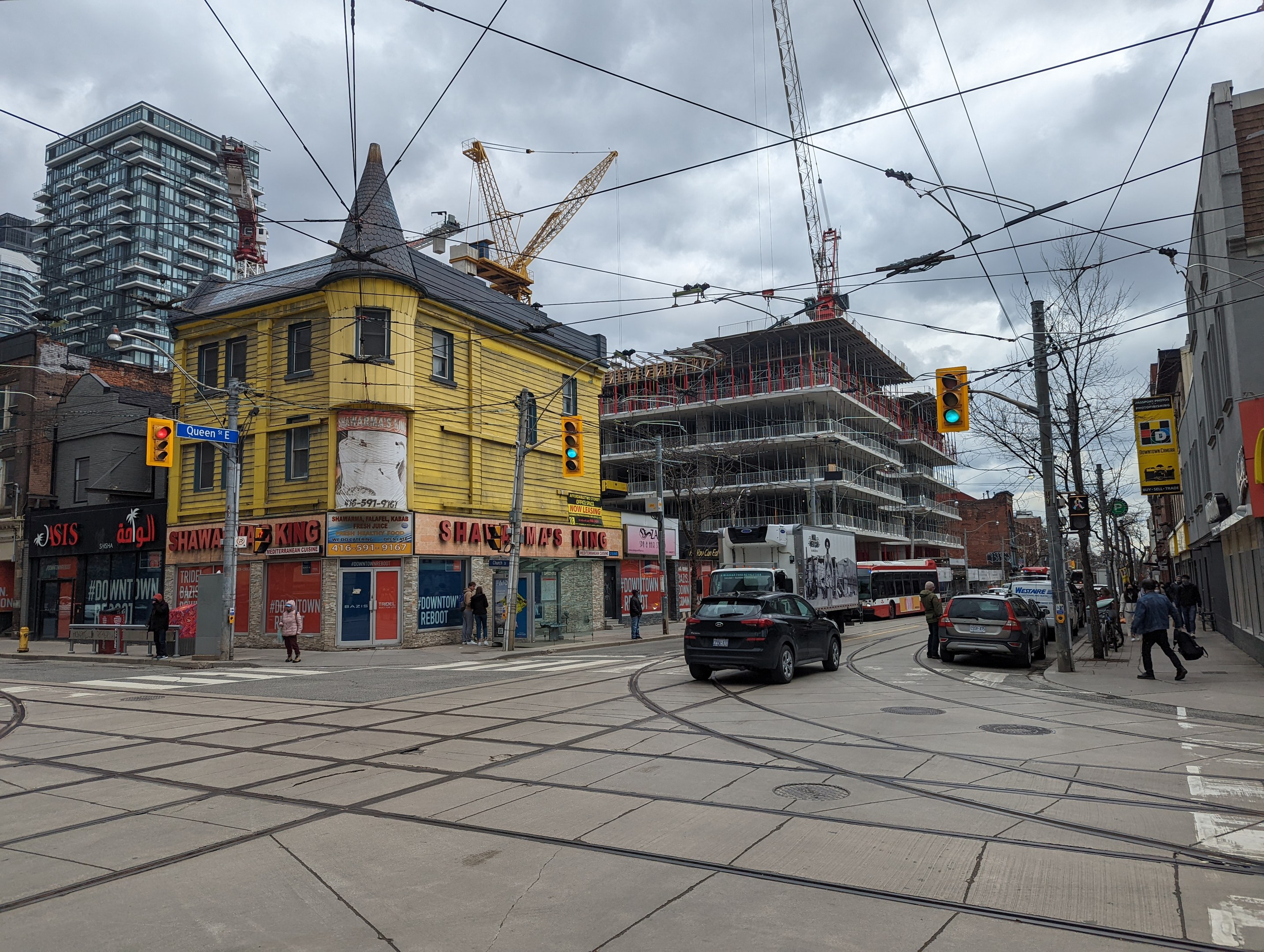 Gelbes Gebäude im Bau im Vordergrund, mit einer Stadtstraße mit Autos, Fußgängern, Ampeln, Strommasten, Gebäuden, Schildern, Bäumen, Kränen und einem bewölkten Himmel im Hintergrund.