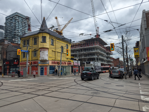 Gelbes Gebäude im Bau im Vordergrund, mit einer Stadtstraße mit Autos, Fußgängern, Ampeln, Strommasten, Gebäuden, Schildern, Bäumen, Kränen und einem bewölkten Himmel im Hintergrund.