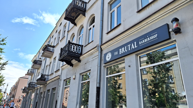 Eine Straßenansicht eines Friseurladens in Berlin, Deutschland, mit Gebäuden, die Fenster, Geländer, Balkone und Namensschilder aufweisen, Bäumen, Laternenpfählen und Menschen, die auf der Straße unter einem bewölkten Himmel gehen.