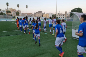Eine Gruppe von Frauen, die auf einem Feld zwischen Gebäuden, Bäumen und Pfählen unter einem klaren blauen Himmel Fußball spielt, mit einem Torpfosten und Netz auf der rechten Seite.