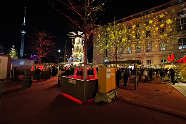 Ein geschäftiger Weihnachtsmarkt in Berlin, Deutschland, mit Menschen um beleuchtete und festlich geschmückte Stände, Bäume, Gebäude, Laternenmasten und einen Turm im Hintergrund unter einem dunklen Himmel.