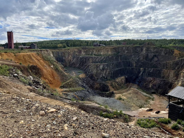 Großer Tagebau auf einem Schmutzfeld umgeben von Bäumen, Gebäuden und einem Schuppen, mit einem bewölkten Himmel und einem Boden voller Steine und Pflanzen.