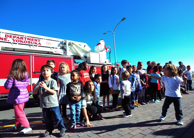 Eine Gruppe von Kindern vor einem Feuerlöschfahrzeug auf einer Straße mit Laternenmasten, Bäumen und einem klaren blauen Himmel, das Feuerlöschfahrzeug wahrscheinlich Teil der Weihnachtsparade der Yokosuka-Feuerwehr.