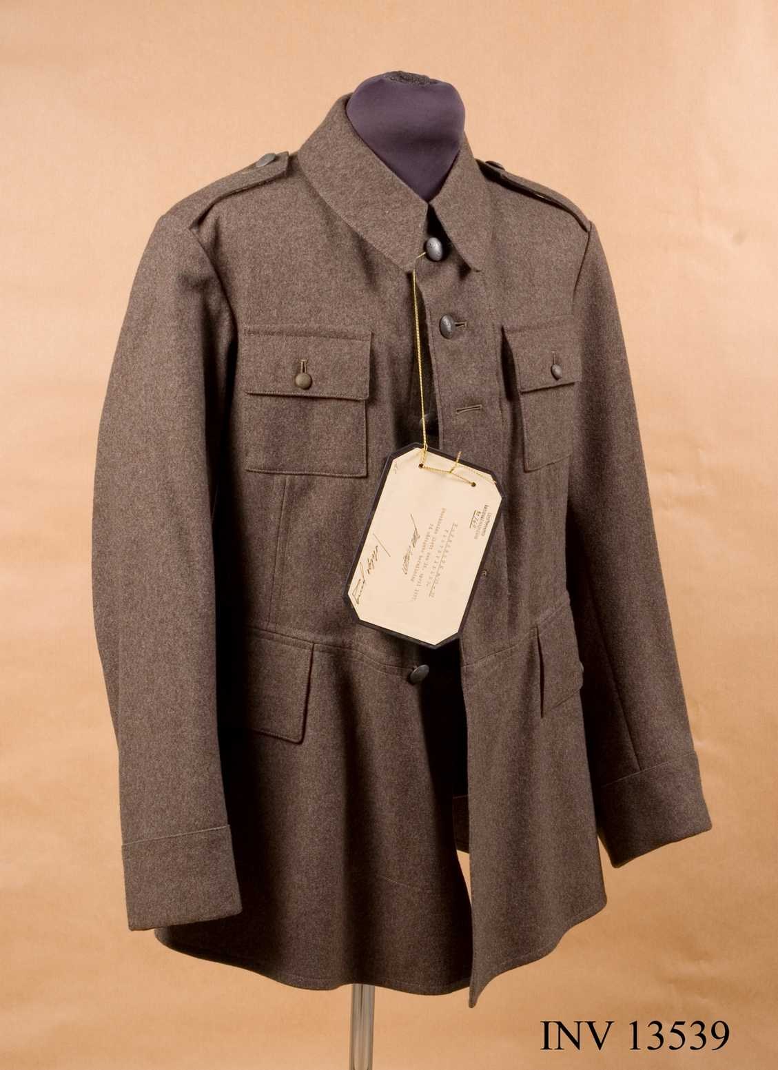 Luftwaffen-Offiziersjacke der deutschen Wehrmacht mit angebrachtem Schild, das auf einer Schaufensterpuppe gegen einen cremefarbenen Hintergrund gestellt ist, mit Text unten.