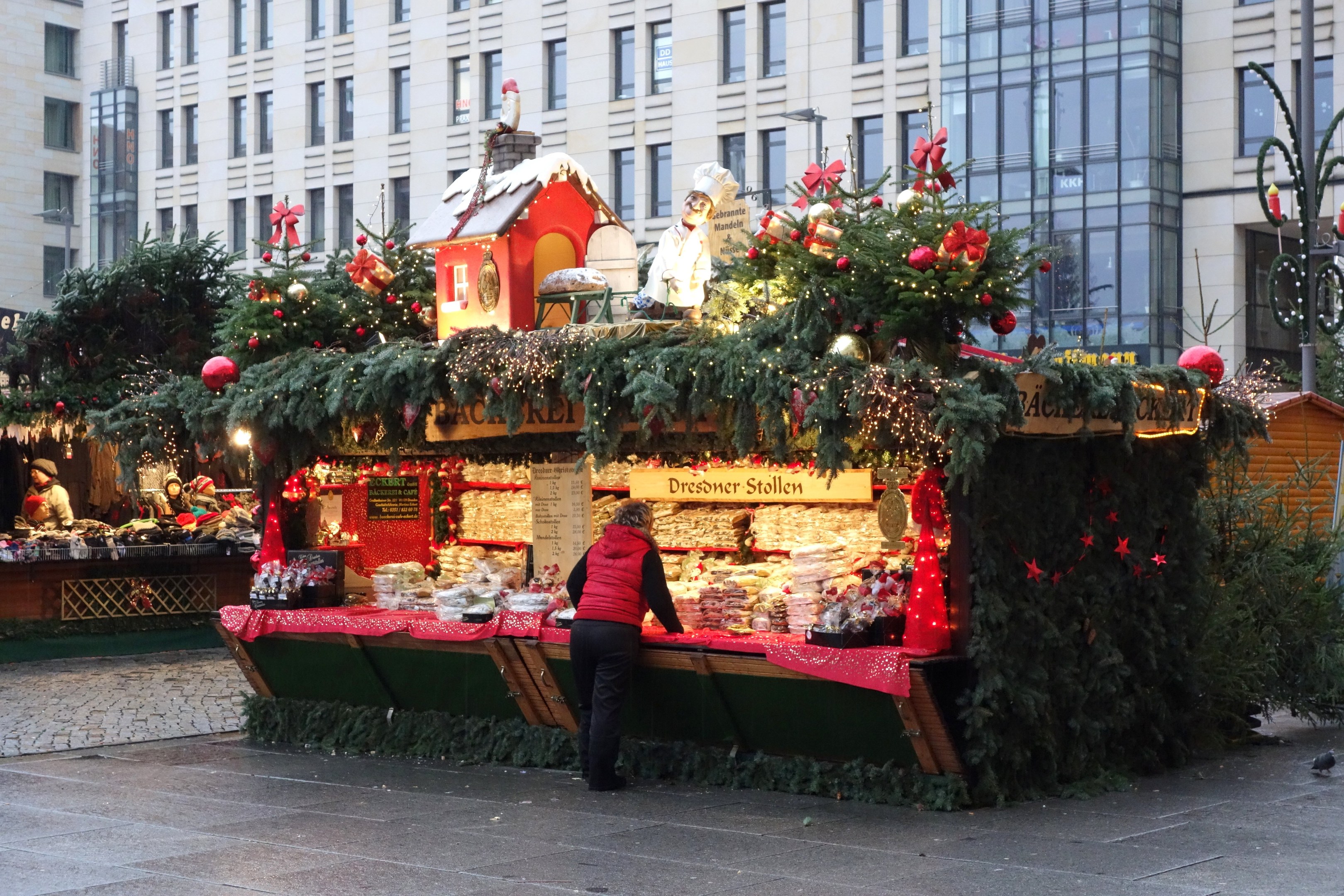 Eine Person steht vor einem Weihnachtsmarktstand, der mit Lichtern, Tannenbäumen und Schmuck geschmückt ist, in einem Stadtplatz mit Gebäuden und einer Straßenlaterne im Hintergrund.