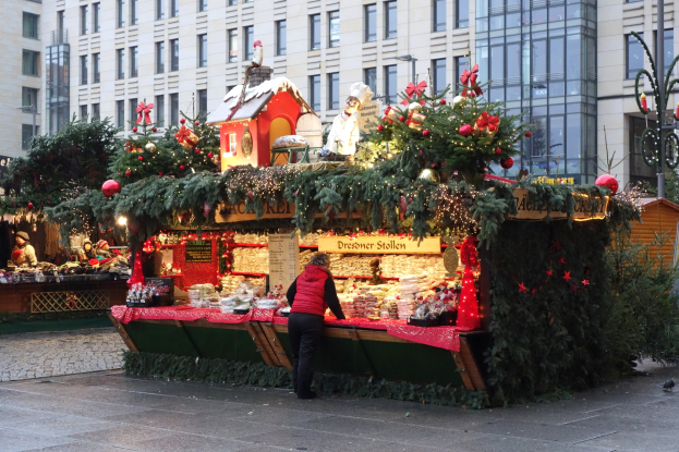 Eine Person steht vor einem Weihnachtsmarktstand, der mit Lichtern, Tannenbäumen und Schmuck geschmückt ist, in einem Stadtplatz mit Gebäuden und einer Straßenlaterne im Hintergrund.