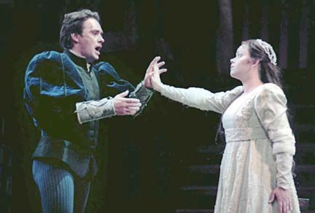 Ein Mann in einem schwarzen Anzug und weißem Hemd und eine Frau in einem langen Kleid stehen auf einer Bühne und sehen ernst aus, wobei der Mann ein Objekt hält und eine Treppe im Hintergrund zu sehen ist.