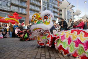Vibrantes chinesisches Neujahrsfest in Amsterdam mit Löwen tanzen im Vordergrund und einer Zuschauermenge, einige halten Kameras, vor einem Hintergrund aus Gebäuden, Laternenmasten und einem klaren blauen Himmel.