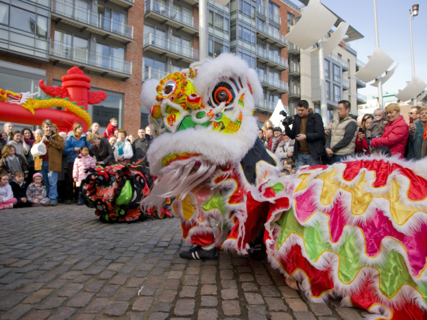 Vibrantes chinesisches Neujahrsfest in Amsterdam mit Löwen tanzen im Vordergrund und einer Zuschauermenge, einige halten Kameras, vor einem Hintergrund aus Gebäuden, Laternenmasten und einem klaren blauen Himmel.