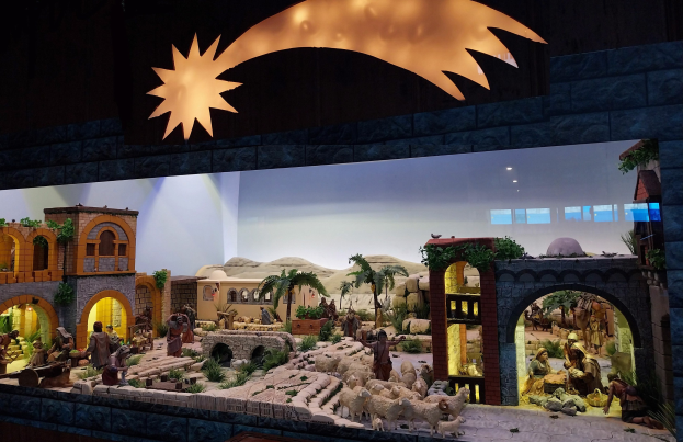 Eine Krippenausstellung im Museum, die Menschen, Tiere, Häuser, Bäume und andere Gegenstände zeigt, mit einer Wand im Hintergrund und einer Lampe, die die festliche Anordnung beleuchtet.