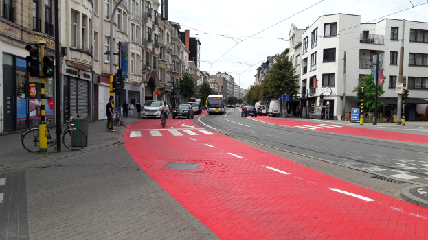 Rote Fahrradspur auf einer Stadtstraße mit Fahrzeugen, Fußgängern, Gebäuden, Bäumen und einem klaren blauen Himmel.