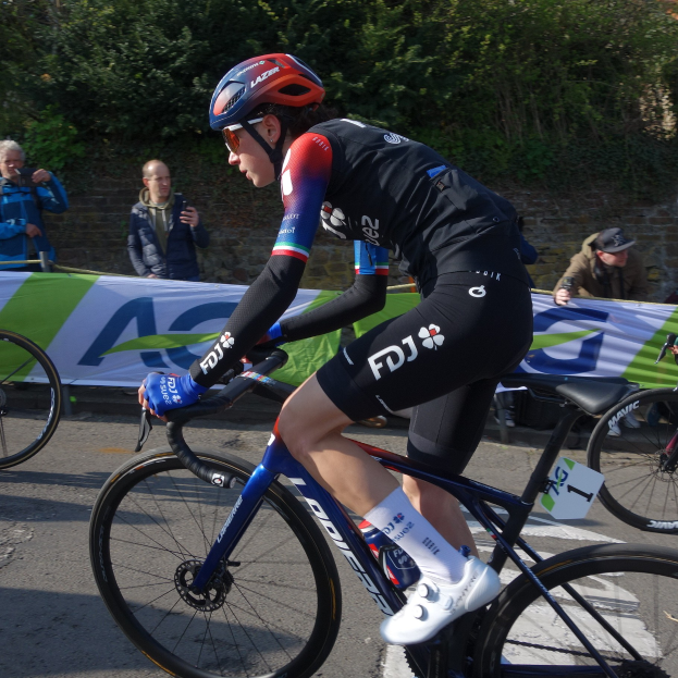 Eine professionelle Radsportlerin, Katie Archibald, fährt mit Helm, Brille, Handschuhen und Schuhen auf einem Fahrrad eine Straße entlang, neben einer Menschenmenge, mit einem Banner, einer Wand, Bäumen und einem Gebäude im Hintergrund.