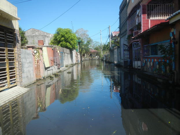Ein schmaler Kanal in einer Stadt, gesäumt von Gebäuden auf beiden Seiten, mit ruhigem, klarem Wasser, das Bäume, Pfähle, Drähte und den Himmel spiegelt.