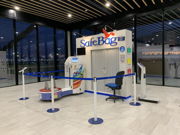 Sicherheitskontrolle am Flughafen mit Stuhl, Absperrgittern, Maschinen, Bildschirm, bedruckter Tafel, Glaswänden, Deckenleuchten und sichtbarem Boden.