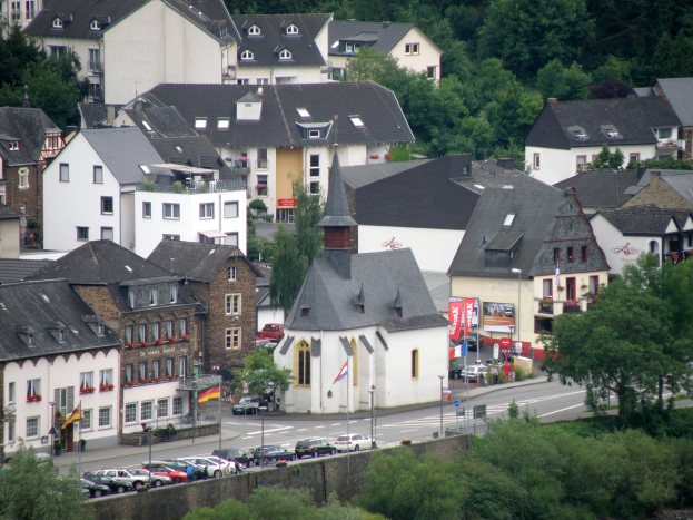 Eine kleine Stadt mit zahlreichen Gebäuden, Fahrzeugen, Bäumen und einer prominenten Kirche in der Mitte, mit Pflanzen und Wasser am unteren Bildrand.