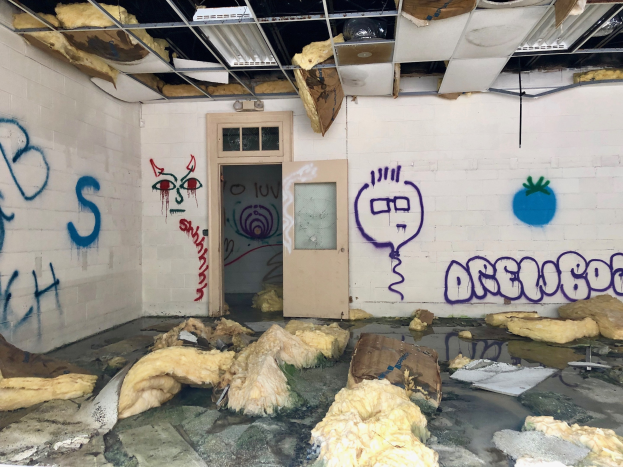 Ein Raum mit Graffiti an den Wänden, eine Tür im Hintergrund, Deckenleuchten und verschiedene Gegenstände auf dem Boden, die tragische Ereignisse im Detroit-Bereich symbolisieren.