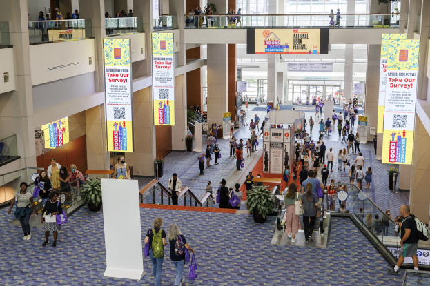 Eine große Menschenmenge bewegt sich durch ein großes Gebäude mit Bannern, Topfpflanzen, Rolltreppen, Geländern, Säulen, bedruckten Tafeln, Glastüren und anderen Elementen, einige tragen Taschen, was auf die Teilnahme an der Nationalen Buchmesse hinweist.