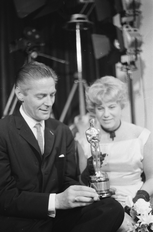 Ein Mann in einem schwarzen Blazer, weißem Hemd und Krawatte hält eine Oscar-Statue, während er neben einer Frau in Abendgarderobe sitzt, die einen Blumenstrauß hält, mit Lichtern im Hintergrund.