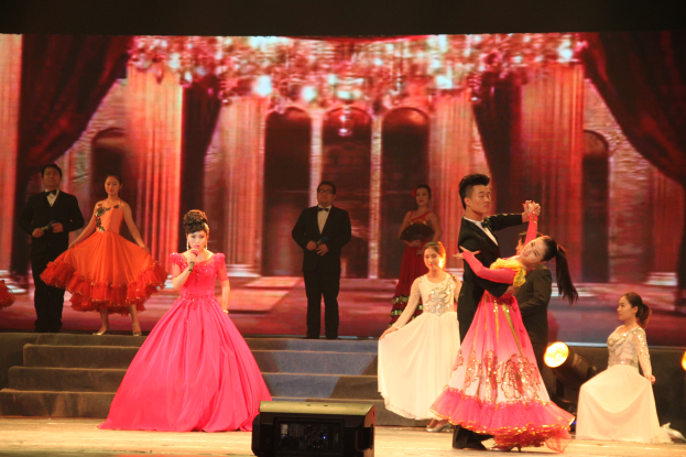 Eine Gruppe von Menschen auf einer Bühne mit Stufen und einem Hintergrundbild, beleuchtet von Scheinwerfern und mit einem Lautsprecher unten, was eine Tanzvorstellung vermuten lässt.