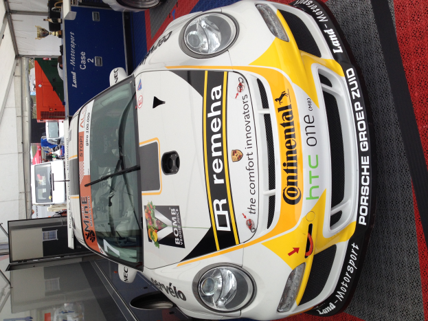 Ein weiß-gelber Porsche 911 GT3 RSR Rennauto in einer Garage geparkt, umgeben von anderen Fahrzeugen, Menschen und diversen Gegenständen.