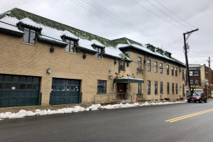 Großes Backsteingebäude mit grüner Dachfläche an einer Straßenkreuzung, identifiziert als ehemalige Feuerwache St. Louis, umgeben von Fahrzeugen, Schnee, Strommasten, Bäumen und einem bewölkten Himmel.