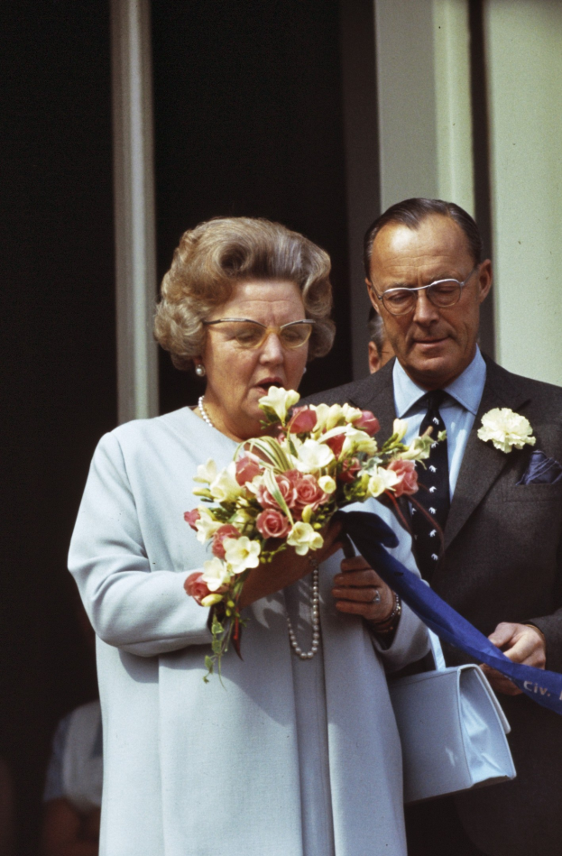 Königin Elizabeth II und Prinz Philip, Herzog von Edinburgh, stehen nebeneinander; sie trägt ein blaues Kleid mit einem Bouquet und er trägt eine Brille und hält ein Objekt, mit anderen und einer Wand im Hintergrund.