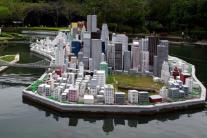 Ein Miniaturstadtmodell, das auf Wasser schwimmt, mit Gebäuden, Grünflächen, einer Brücke, Menschen und Bäumen vor einem Himmel als Hintergrund.