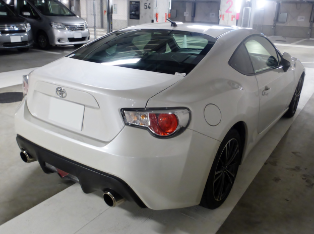 Ein weißer Sportwagen steht in einer Parkgarage neben anderen Fahrzeugen, mit Deckenlampen, die den Raum beleuchten und einer Wand im Hintergrund.