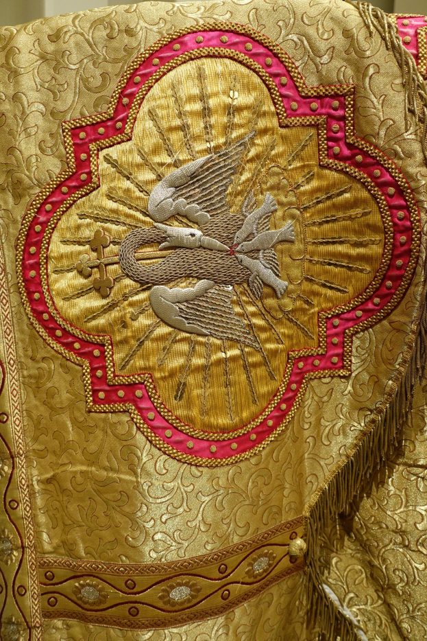 Nahaufnahme eines Priestergewänders mit aufwendiger goldener und roter Stickerei und einem luxuriösen, krönenden Design vor einem neutralen Hintergrund.
