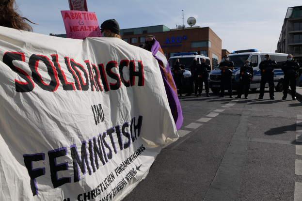 Eine Gruppe von Menschen marschiert auf einer Straße, hält ein Banner mit der Aufschrift "Solidarität und Feminismus" und fährt parkende Fahrzeuge auf der Seite und Gebäude im Hintergrund unter einem klaren blauen Himmel.