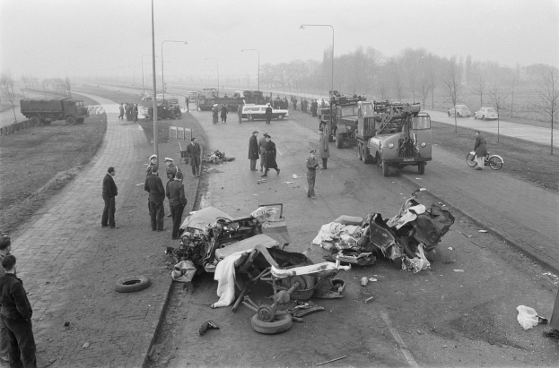 Schwarz-weiß-Szene eines Autounfalls am Straßenrand mit mehreren Fahrzeugen und einer Gruppe von Menschen, Laternen, Bäumen und Himmel im Hintergrund.
