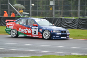 Ein Auto rennt auf einer Strecke mit Text und Zahlen darauf, umgeben von Gras, mit einem Maschendrahtzaun, der mit schwarzem Stoff bedeckt ist, Menschen in orangefarbenen Jacken mit Schirmen und Bäumen im Hintergrund.