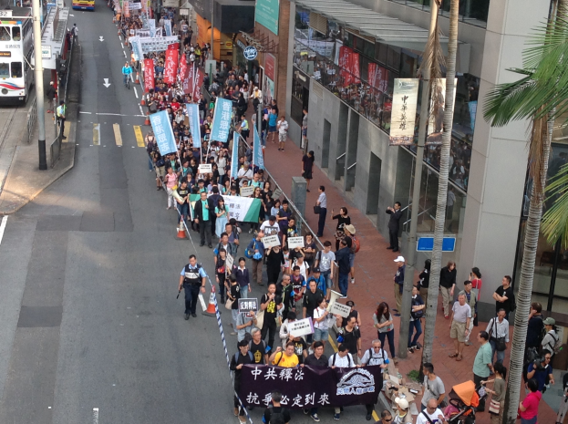 Eine große Protestdemonstration in Hong Kong mit Menschen, die Schilder und Plakate tragen, flankiert von Bäumen, Gebäuden, Fahrzeugen und Straßenschildern.
