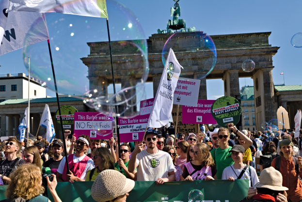 Eine Menschenmenge steht vor dem Brandenburger Tor in Berlin, Deutschland, wobei einige Mützen und Schutzbrillen tragen und Fahnen und Plakate halten, einschließlich eines im Vordergrund, vor einem Hintergrund aus Gebäuden, einem statuebekrönten Bogen und einem Himmel mit Blasen.