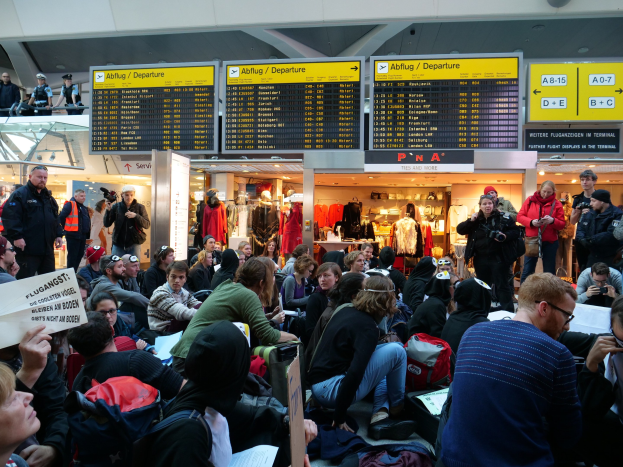 Eine große Gruppe von Menschen, die auf Stühlen und Stehplätzen in einem Flughafen während einer Demonstration sitzen, mit Schildern, Puppen und Deckenlampen im Hintergrund.