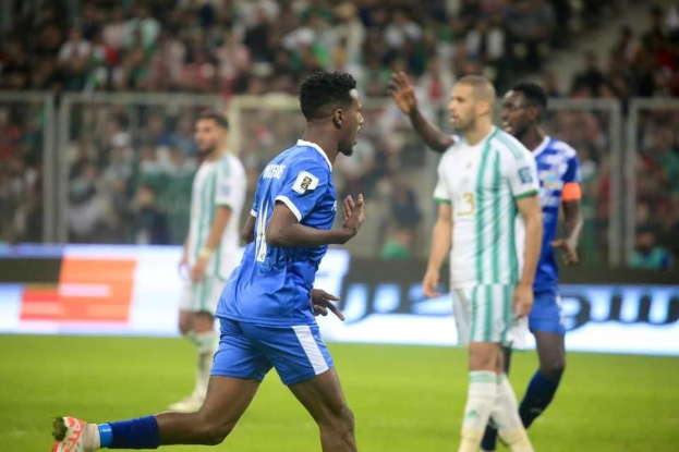 Ein Fussballspieler in einem blauen und weissen Trikot feiert mit erhobenen Armen, umgeben von einer Gruppe in weissen und grünen Trikots, vor einem Hintergrund mit einem weissen und blauen Brett und Geländer.