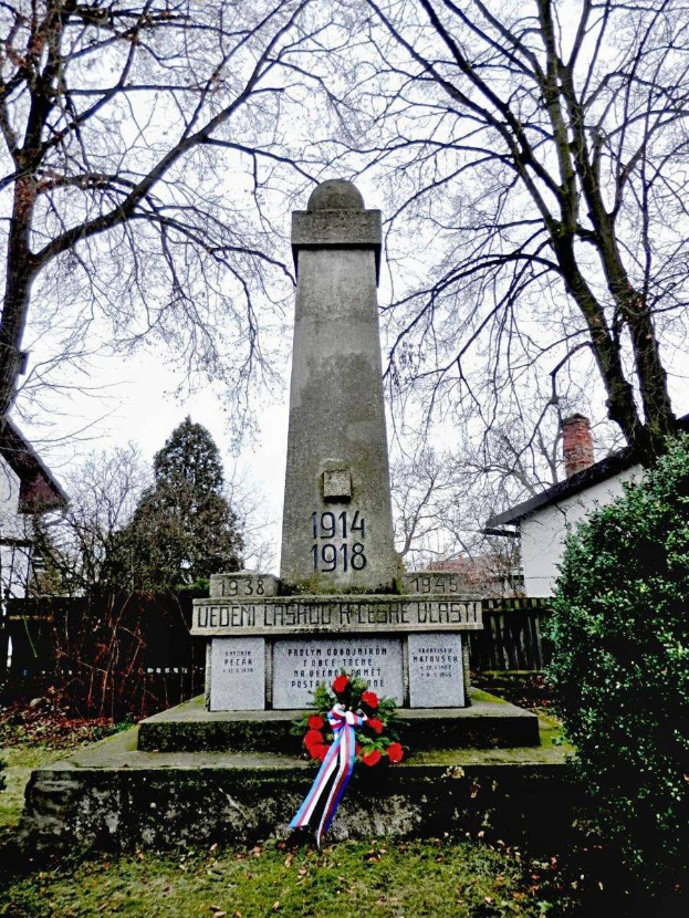 Ein Weltkriegsgedenkmal auf einem Friedhof, geschmückt mit einem Kränzchen, umgeben von Gras und trockenen Blättern, mit Pflanzen, Bäumen, Häusern und dem Himmel im Hintergrund.