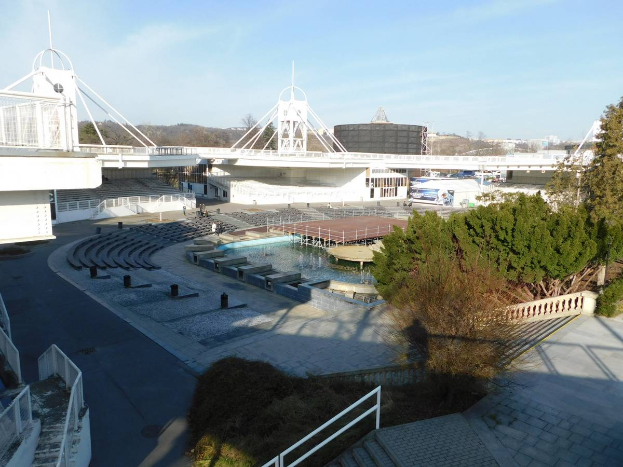 Ein Blick auf das Olympische Stadion von einem hohen Standpunkt aus, mit verschiedenen Objekten und Grünflächen im Vordergrund und einem bewölkten Himmel im Hintergrund.