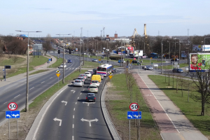Eine belebte Stadtstraße mit zahlreichen Autos, Bussen und Straßenlaternen, umgeben von Schildern, Lichtmasten, Bäumen, Gras, Gebäuden, Türmen und einem bewölkten Himmel.