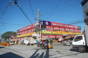Eine belebte Straßenszene mit einem Lastwagen, mehreren Fahrzeugen, Fußgängern, einem Strommast mit Kabeln, Bäumen und einem klaren blauen Himmel im Hintergrund.
