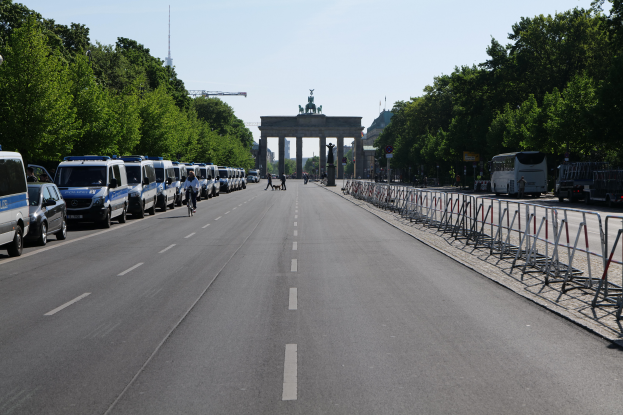 Eine Reihe von Polizeiwagen, die auf der Seite einer Straße vor dem Brandenburg-Tor in Berlin, Deutschland geparkt sind, mit Menschen auf Fahrrädern und in der Nähe stehenden Menschen, Barrieren, Bäumen und einem Bogen mit Statuen im Hintergrund.