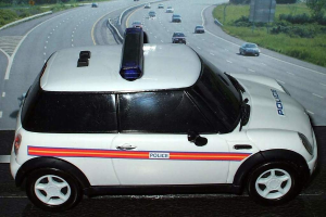 Weißes Polizeiauto mit "Polizei"-Aufschrift auf einer Straße vor der Stadt, umgeben von fahrendem Verkehr, Gras und Bäumen auf beiden Seiten.